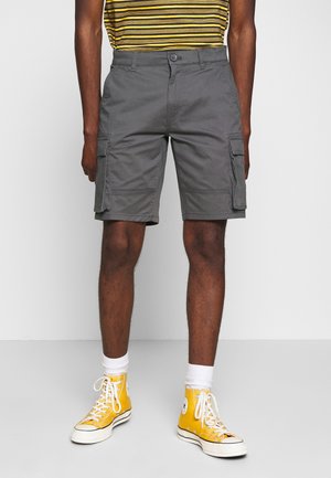 Homme portant un short cargo gris, des baskets montantes jaunes, des chaussettes blanches et une chemise rayée jaune et noire, debout sur un sol gris.