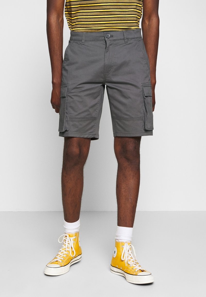 Homme portant un short cargo gris, des baskets montantes jaunes, des chaussettes blanches et une chemise rayée jaune et noire, debout sur un sol gris.
