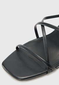 Sandália preta com ponta quadrada, finas tiras entrelaçadas e uma textura de couro liso. O design é minimalista e elegante.