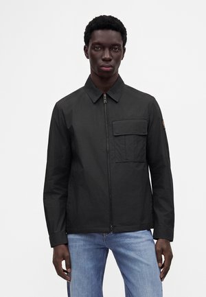 Mann trägt eine schwarze langärmelige Jacke mit Reißverschluss und Brusttasche sowie hellblaue Jeans, steht vor einem schlichten weißen Hintergrund.