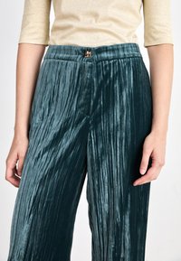 Pantalons en velours plissé teal avec une taille haute et une fermeture à agrafe en métal. Texture lisse avec des lignes verticales, offrant un drapé fluide.