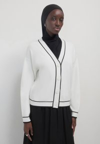 Donna che indossa un hijab nero, un abito nero e un cardigan bianco con bottoni e rifiniture nere, in piedi contro uno sfondo chiaro e semplice.