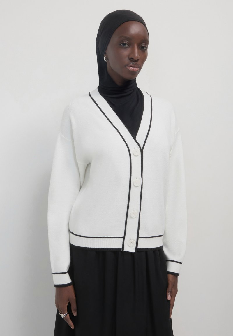 Donna che indossa un hijab nero, un abito nero e un cardigan bianco con bottoni e rifiniture nere, in piedi contro uno sfondo chiaro e semplice.