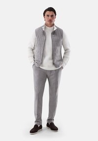 Graue gesteppte Weste über einem weißen, strukturierten Strickpullover, kombiniert mit hellgrauen, maßgeschneiderten Hosen und dunklen Wildlederloafern. Schlichte, klare Linien.