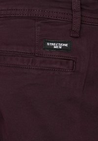 Burgundy-housut, joissa on sileä pinta, varustettuna takataskulla ja mustalla tarralla, jossa lukee "STREETONE MEN." Saumojen varrella on ommeltuja yksityiskohtia.