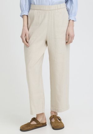 Nő, bézs színű, széles szárú, rugalmas derékpántos nadrágban, barna szarvasbőr, csatos, bebújós cipővel.