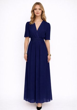 Vrouw staand, gekleed in een marineblauwe maxi-jurk met V-hals, korte mouwen en geplooide taille, gecombineerd met zwarte hakken en minimalistische sieraden.