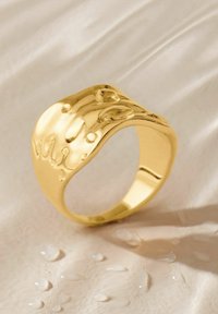 Goldring mit wellenförmigem Design, das mit erhabenem Detail und runden Formen verziert ist. Glatte Oberfläche mit Wassertropfen auf einem hellen Hintergrund.