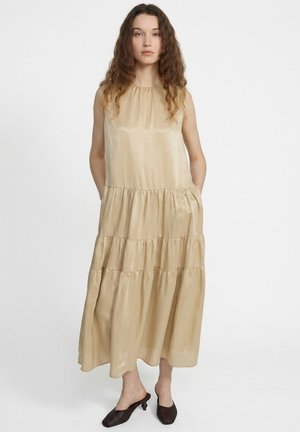 Femme aux longs cheveux bouclés portant une robe beige sans manches à volants et des mules noires à talons, debout devant un fond blanc uni.