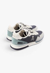 Pár nízkých tenisek s béžovými, námořnickými a tyrkysovými panely, zavazováním na šňůrky, silnými bílými podrážkami a logem lebky a zkřížených kostí na boku.