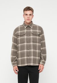 Bruine geruite shirt gemaakt van stof met een gestructureerde afwerking. Heeft een knoopsluiting aan de voorkant, twee borstzakken en een kraag.