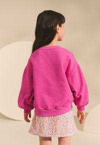 Hanorac fucsia cu mâneci lungi și bufante, cu tivuri dantelate; asortat cu o fustă roz pal, cu paiete.