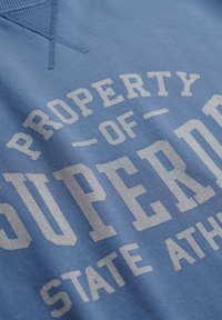 Sudadera azul con texto blanco que dice "PROPIEDAD DE SUPERDRY STATE ATH." Presenta un cuello redondo texturizado y una tela suave de algodón.