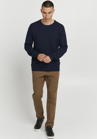 Pulover texturat bleu marin cu mâneci lungi, decolteu rotund și manșete cu nervuri, asortat cu pantaloni eleganți maro și adidași negri.