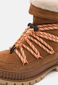 Brune semskede støvler med en robust, brun gummisål. Har oransje-svart mønstrede snøringer og en fleecefôret krage.