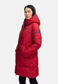 Woman wearing a long red padded coat with hood, black zipper, and patch on left sleeve, standing with hands in pockets, facing slightly left.
