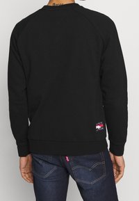 Czarna bluza z okrągłym dekoltem i raglanowymi rękawami. Charakterystyczna metka na dole. Noszona z ciemnymi dżinsami. Gładka tekstura tkaniny.