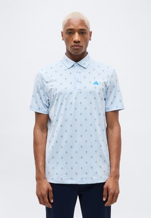 Lühemate heledate juustega mees, kes kannab helesinist mustriga polosärki ja tumedaid pükse, seisab valge ühevärvilise tausta ees.