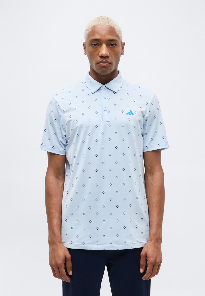 Άνδρας με κοντά ξανθά μαλλιά, ντυμένος με ανοιχτό μπλε πόλο με σχέδια και σκούρο παντελόνι, στέκεται μπροστά σε απλό λευκό φόντο.