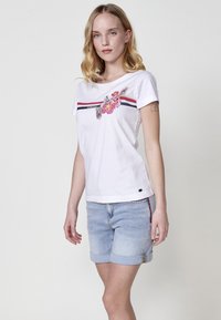 Camiseta de algodón blanca con un diseño floral en tonos rosa y verde, y rayas rojas y azul marino en el pecho. Combinada con pantalones cortos de mezclilla claros.
