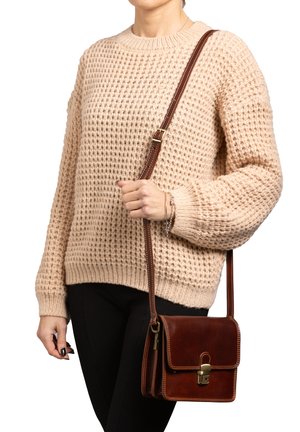 Braune Leder-Crossbody-Tasche mit Klappe, goldenen Beschlägen und weißen Nähten, kombiniert mit einem cremefarbenen Strickpullover und schwarzen Hosen.