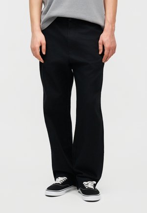 Egy személy laza fekete nadrágot, fekete-fehér tornacipőt és szürke inget visel, miközben egy egyszínű világos háttér előtt áll.