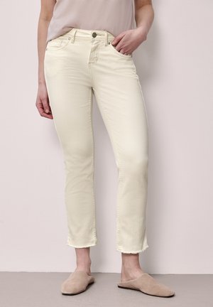 Kvinde iført cremefarvede, tætsiddende jeans med flossede kanter, afslappede beige slip-on sko og en lysebeige top, stående op ad en bleg væg.