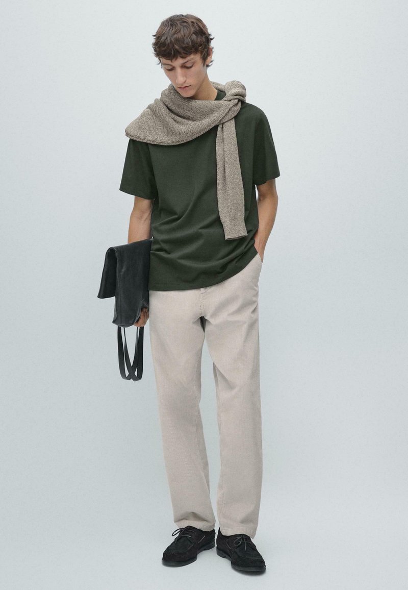 Maglietta verde scuro, pantaloni di velluto a coste beige, sciarpa grigia drappeggiata sulle spalle, borsa nera e scarpe in camoscio nero. Sfondo neutro.