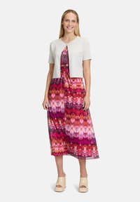 Lichtgrijs kortemouwen cardigan over een kleurrijke, patroonrijke mouwloze maxidress in tinten roze en rood, met een knoopdetail in de taille.