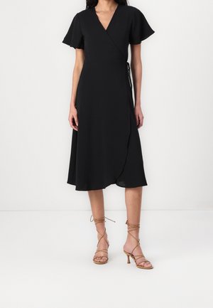 Kobieta w czarnej sukience kopertowej z krótkimi rozkloszowanymi rękawami i beżowych wysokich sandałach na sznurowadła, stojąca na tle białego tła.