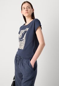 Navy blue cotton t-shirt with a white graphic design featuring leaves and waves, round neckline, and short sleeves. Paired with loose trousers.