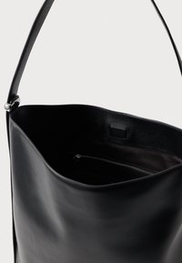 Bolsa tote de cuero negro con un exterior suave, parte superior abierta y un bolsillo interior con cremallera. Cuenta con una resistente correa para el hombro.