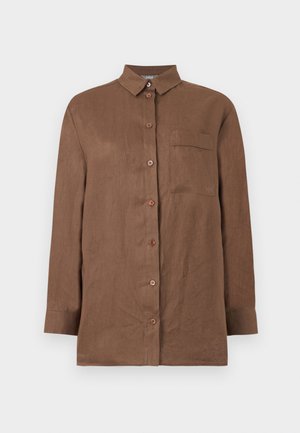 Kobieta stoi ubrana w brązowe spodnie o szerokich nogawkach oraz dopasowaną brązową koszulę zapinaną na guziki na białą koszulkę na ramiączkach, z czarnymi sandałami na obcasie z paskami.