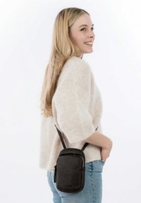 Malá černá kožená crossbody kabelka s texturovaným povrchem, zipovým uzávěrem a nastavitelným popruhem, nošena na lehkém svetru a džínech.