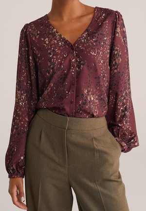 Femme portant un chemisier bordeaux à manches longues avec des taches abstraites et un pantalon taille haute olive, une main dans la poche, fond neutre.