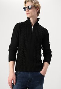 Pull en tricot noir avec un col montant zippé, motifs en relief à torsades, et poignets et ourlet côtelés. Porté avec un jean en denim bleu.