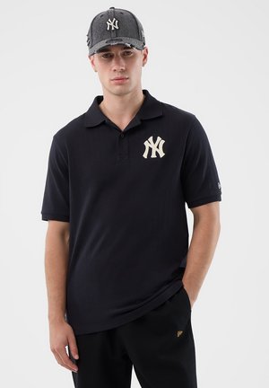 Joven con gorra negra de los New York Yankees, polo negro con el logo de los Yankees y pantalones negros, de pie con una mano en el bolsillo.