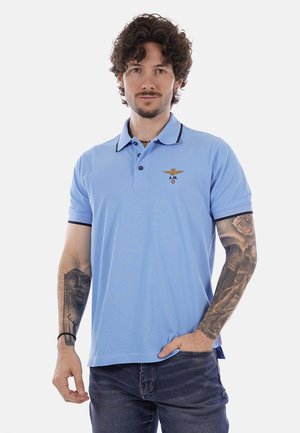 Uomo con capelli ricci che indossa una polo azzurro chiaro con logo ricamato e jeans scuri, mostrando gli avambracci completamente tatuati, in piedi davanti a uno sfondo bianco.