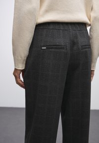 Pantalons noirs à motifs avec un tissu texturé, comportant deux poches arrière et une taille élastique, complétés par un pull de couleur claire.
