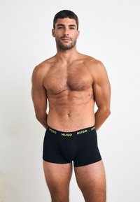 Boxer briefs neri con un logo "HUGO" giallo sulla vita. Il tessuto appare liscio ed elastico, aderente. Il modello è senza camicia.