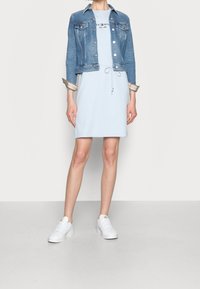 Osoba w jasnoniebieskiej sukience z logo, niebieskiej dżinsowej kurtce i białych trampkach, stojąca na jednolitym jasnym tle.