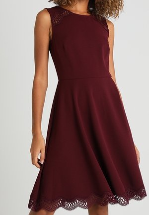 Ärmelloses bordeauxfarbenes Kleid mit Spitzen-Details an Schultern und Saum, tailliertem Bund und ausgestelltem Rock, getragen von einem stehenden Model.