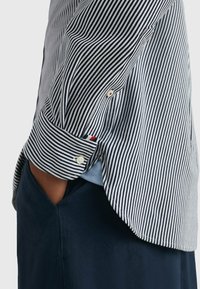 Camisa de botones a rayas en algodón azul marino y blanco. Presenta un puño doblado con acentos contrastantes en rojo y azul y un dobladillo redondeado.