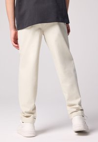 Világos bézs pamut nadrág laza szabással, felhajtott szegéllyel és oldalzsebekkel, fehér sportcipővel semleges háttéren.