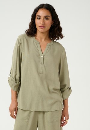 Blouse légère vert clair avec un col en V, trois boutons et des manches retroussées. Texture douce et coupe décontractée avec des détails de couture subtils.