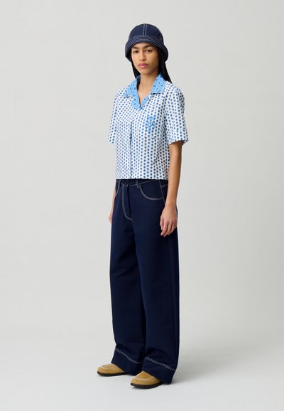 Camicia a maniche corte blu e bianca a pois con colletto, abbinata a pantaloni a gamba larga blu scuro e scarpe gialle con dettagli bianchi.
