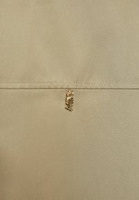 Beige stof met horizontale naad en een kleine gouden hanger die hangt, met gestileerde letters "MW" die onder de naad zijn bevestigd.