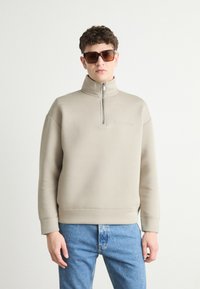 Felpa beige con zip e collo alto, texture morbida e logo ricamato. Abbinata a jeans in denim blu chiaro e occhiali da sole scuri.