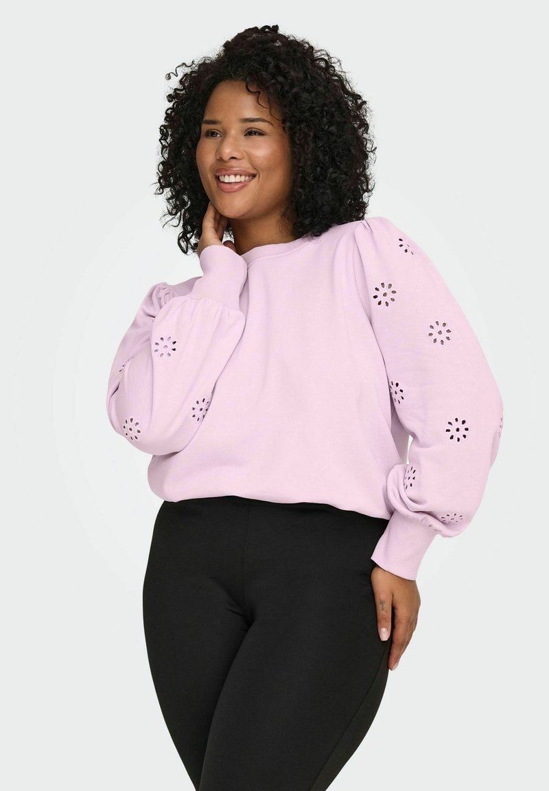 Svetlo roza pulover z napihnjenimi rokavi, okrašen z vzorci okroglih izrezkov; kombiniran s črnimi pajkicami. Gladek teksturni material.