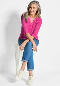 Lebendige pinke Bluse mit Knopfdetails, kombiniert mit hochgekrempelten blauen Jeans und weißen Sneakers, vor einem schlichten Hintergrund.